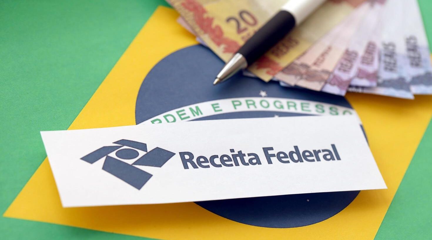 Logotipo da Receita Federal sobre a bandeira do Brasil, com notas de dinheiro e caneta, representando obrigações e regularidade fiscal.